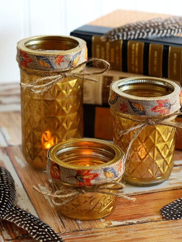 Gold painted quilted jelly jars used as votive holders sitting on a wooden background with feathers and books.