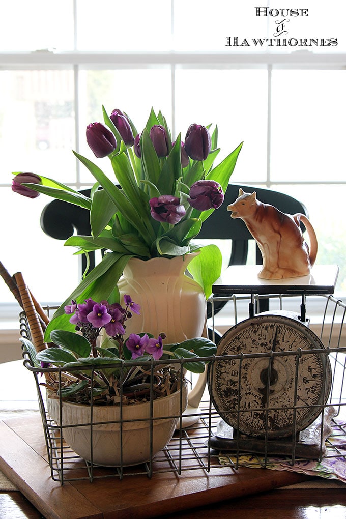 A white vase of tulips and an African violet as part of a rustic farmhouse vignette on a kitchen table.  