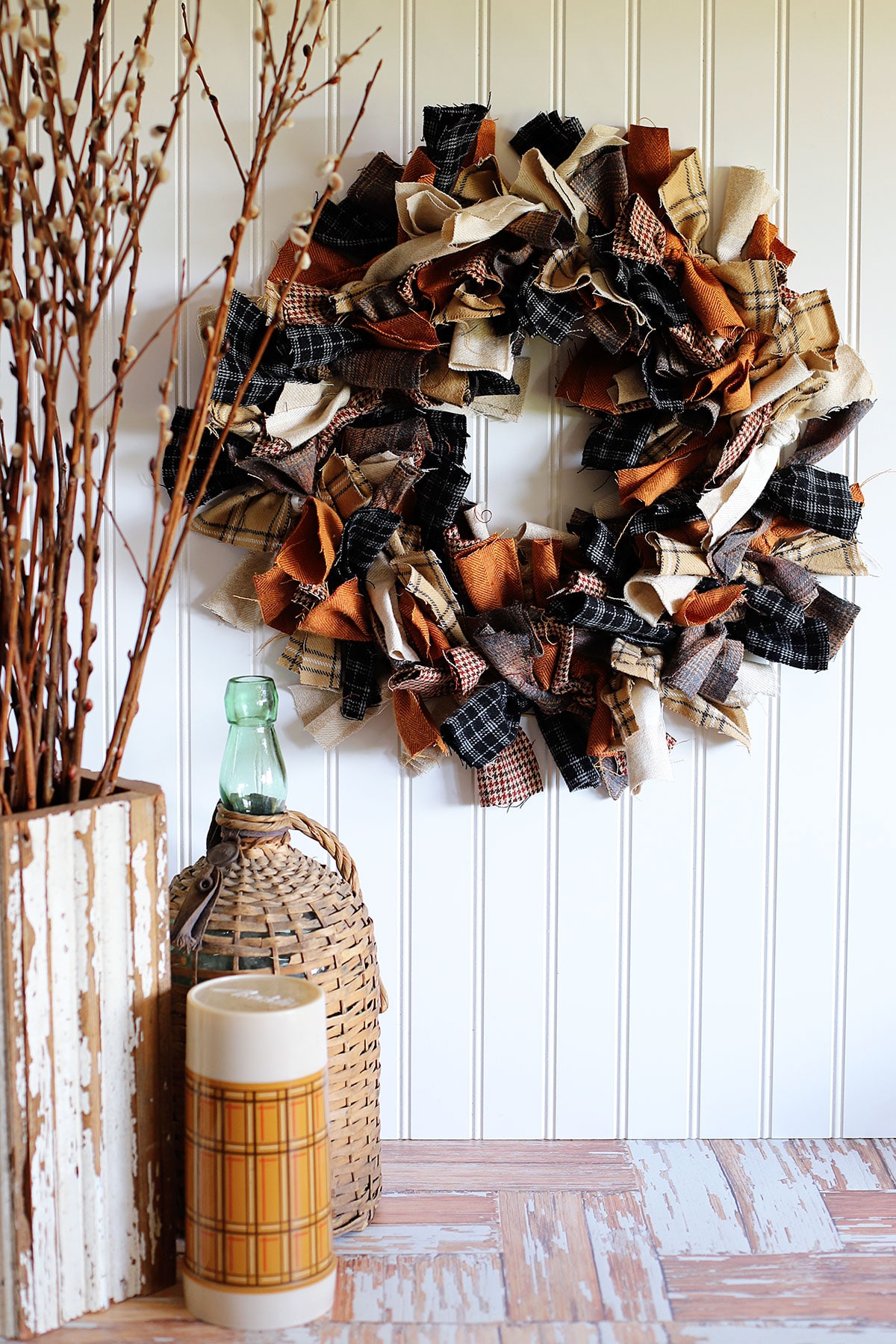 Tutorial for making a rag wreath - showing a rag wreath hanging on the wall next to a wooden vase of twigs, old wicker covered wine bottle and a brown thermos.