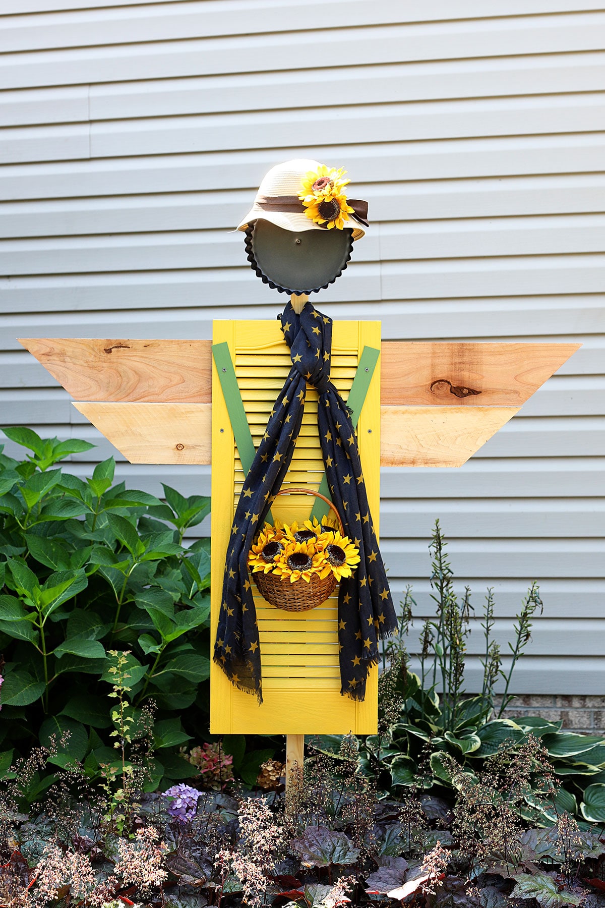 An bright yellow upcycled shutter made into a scarecrow - wearing a scarf and a hat covered in sunflowers and carrying a basket of sunflowers.