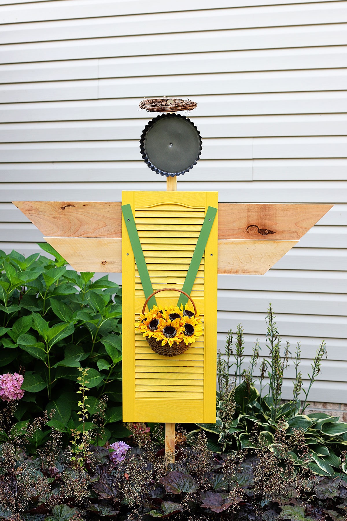 A shutter angel for the garden made with a repurposed shutter from the thrift store. The angel is holding a basket of sunflowers and wearing a halo made from a grapevine wreath.